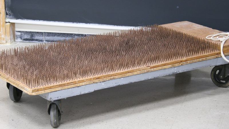 Plywood bed on casters densely packed with upward-pointing nails, with metal handles on both ends