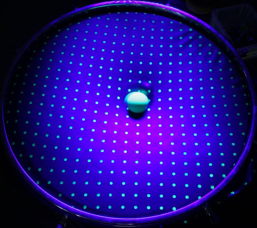 Top-down UV view of neoprene sheet with fluorescent dot grid; a single glowing fluorescent ball sits near center on the relatively flat surface with no visible central mass