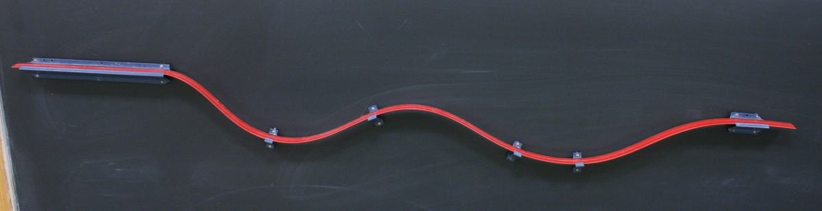 Red flexible track mounted on a blackboard with magnetic clips, shaped in a wavy path with two dips, elevated at the left end and lower at the right