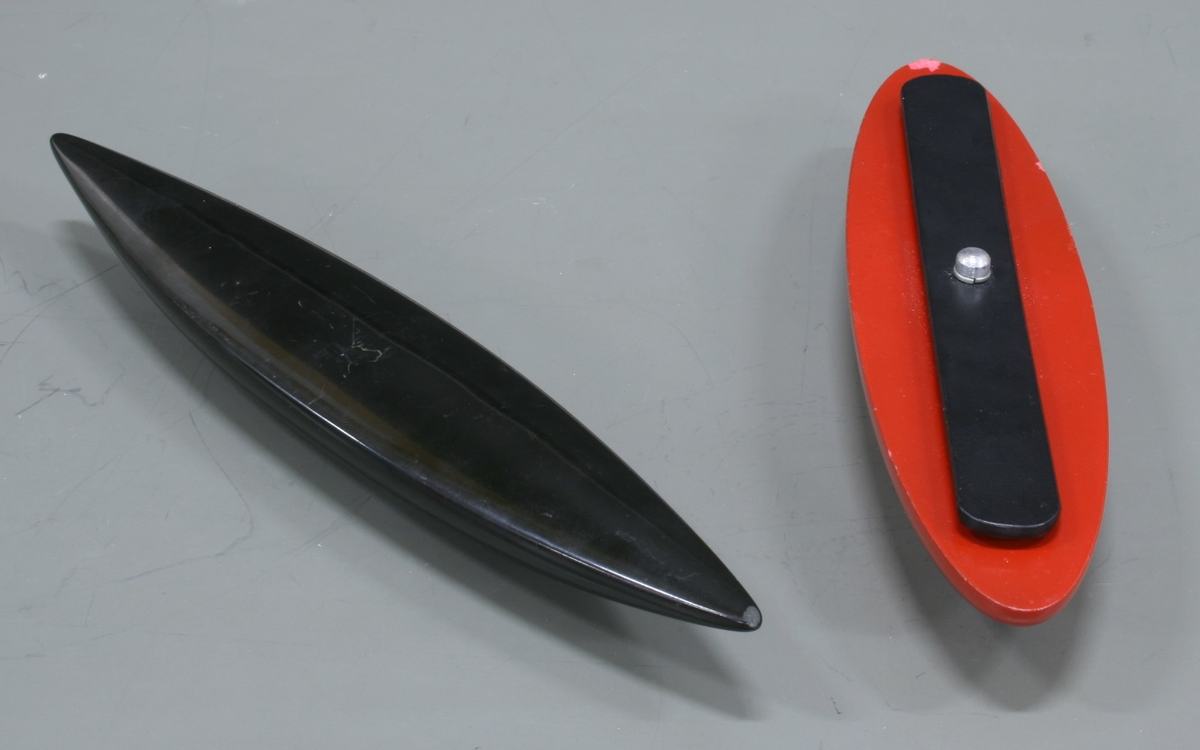 Two rattlebacks on a gray surface: a black elongated boat-shaped rattleback (left) and a shorter red oval rattleback with an adjustable black vane secured by a silver screw (right)