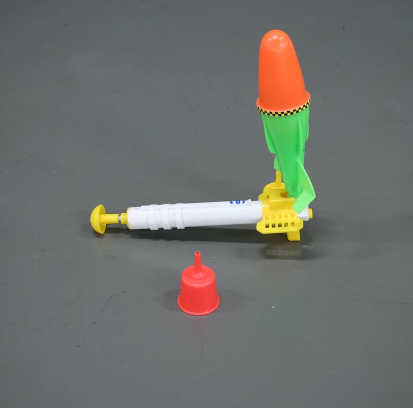 Toy water rocket with green body, orange nose cone, and fins, connected to yellow launch base with white syringe-style pump; red funnel in foreground