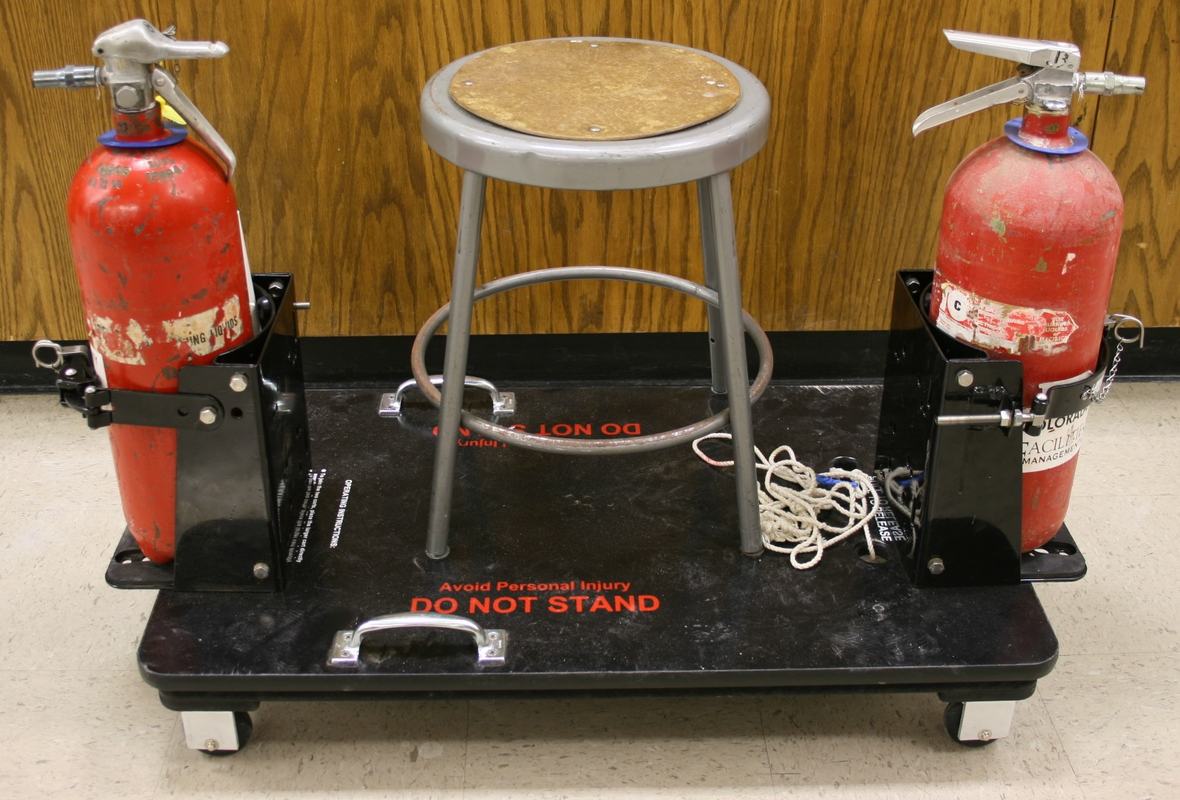 Black wheeled platform on casters with two red CO₂ fire extinguishers in metal brackets, a gray stool bolted to the center of the deck, and 'DO NOT STAND' painted on the deck surface