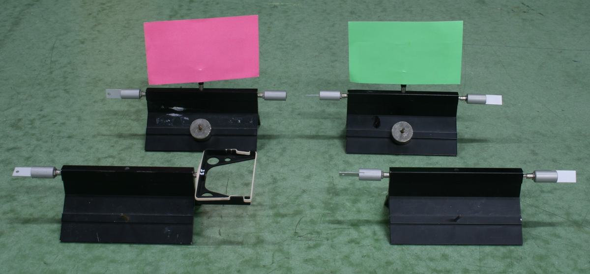 Four black air track gliders arranged in two pairs on a green surface, with pin, wax cup, blade, and bumper attachments on their ends; two gliders have pink and green index card flags for visibility