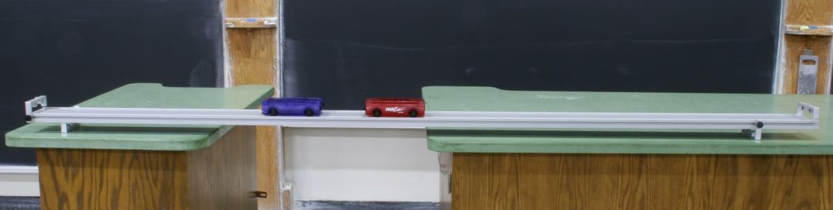 Two PASCO dynamics carts (one blue, one red) on an aluminum dynamics track, set up on a demonstration table in front of a chalkboard