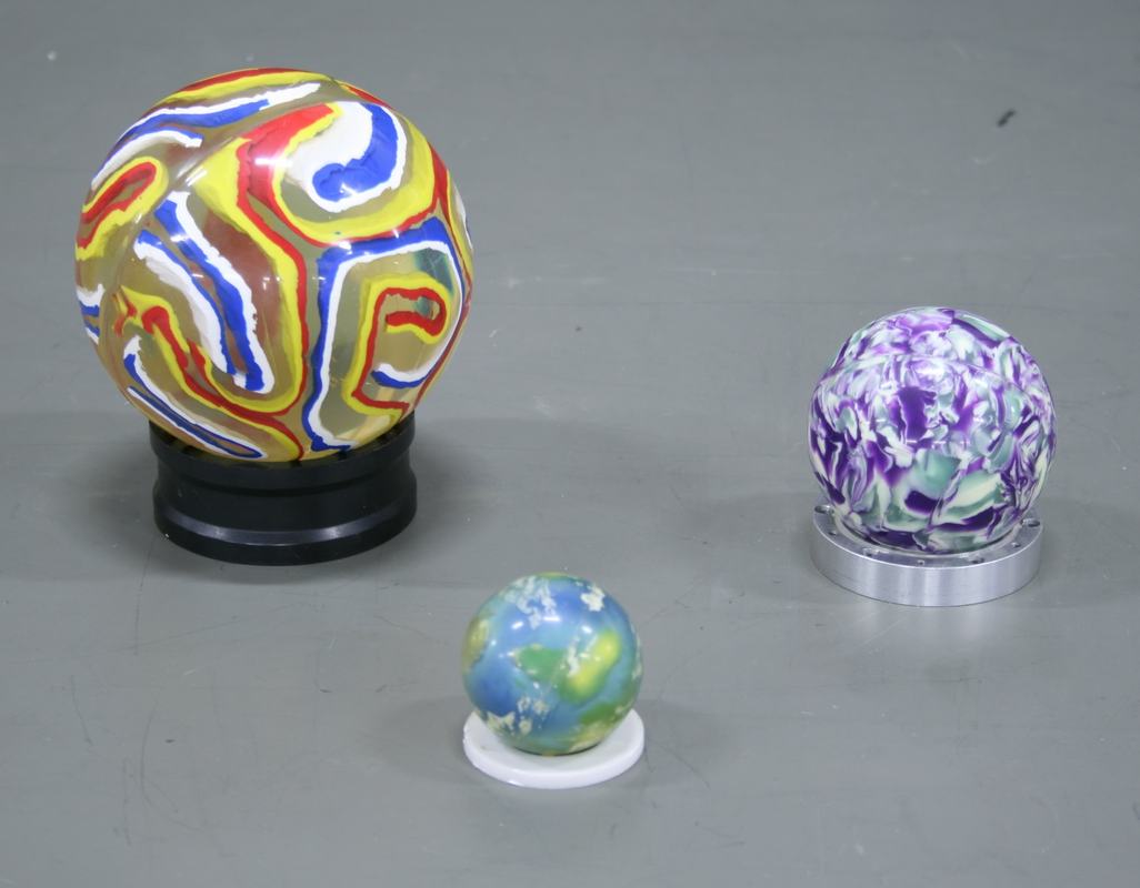 Three superballs of different sizes on ring stands: large multicolored swirled ball (4.5 in.) on black stand at left, small earth-patterned ball (2 in.) on white stand in center, medium purple and white marbled ball (3 in.) on silver stand at right