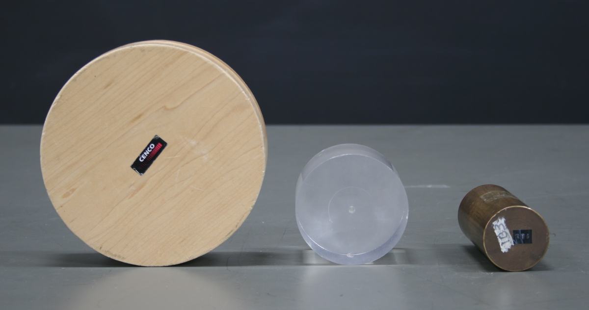 Three objects on a gray surface: large wooden disk with CENCO label (left), medium translucent acrylic disk (center), and small brass cylinder lying on its side (right)