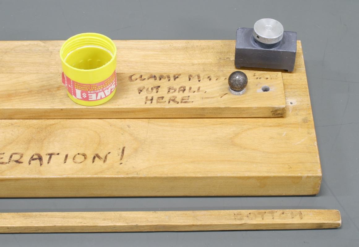 Close-up of base end showing yellow cup, ball bearing in metal cradle, hinge clamp with thumbscrew, and text burned into wood reading "CLAMP" and "PUT BALL HERE"