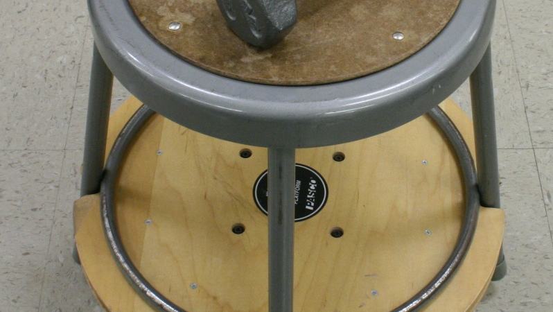 Gray metal stool seated inside a wooden rotating turntable, with two gray dumbbells on the stool seat