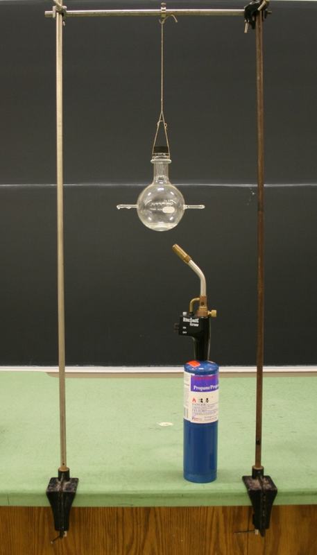 Round-bottom glass flask with two horizontal glass exit tubes, suspended by string from a horizontal rod between two bench clamp stands; a BernzOmatic propane torch sits below on the bench