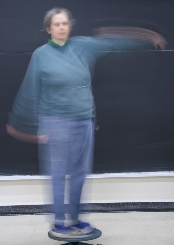 Long-exposure photo showing motion blur as the person swings the hand weight in an arc, with the platform and body visibly rotating in the opposite direction
