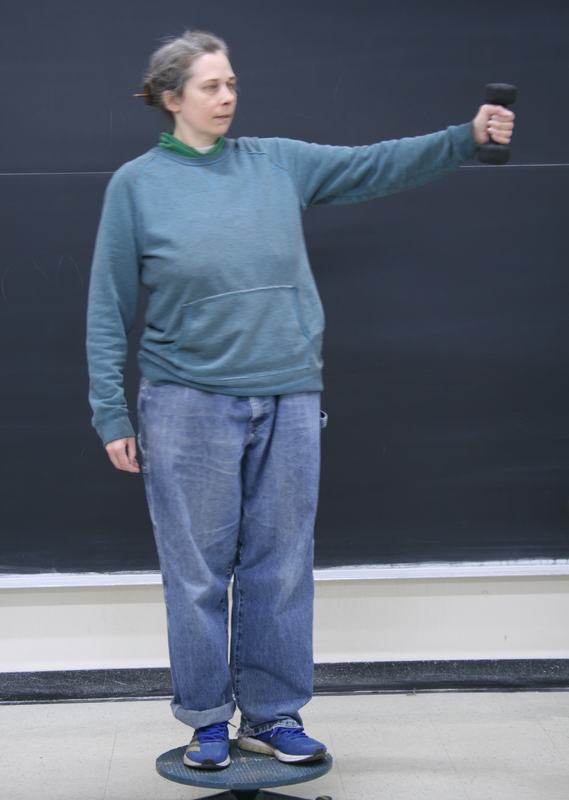 Person standing on a small rotating platform in front of a blackboard, holding a hand weight extended at arm's length