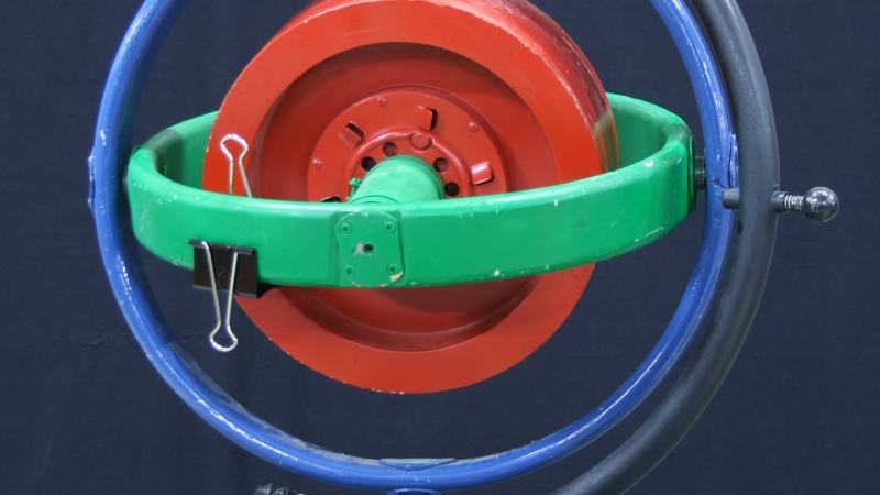 MITAC motorized gyroscope with red rotor disk in green inner gimbal ring and blue outer gimbal ring, mounted on black base with power cord, two wooden blocks with binder clips on table nearby