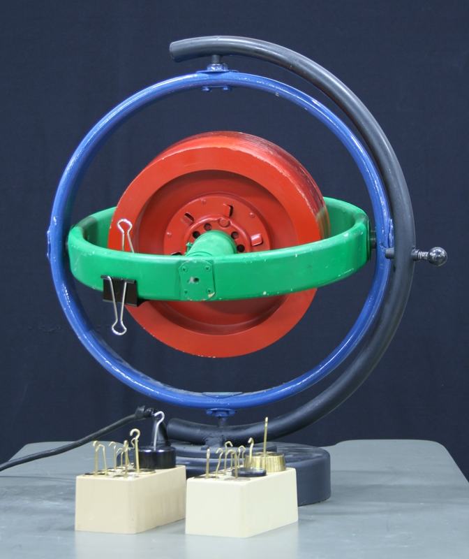 MITAC motorized gyroscope with red rotor disk in green inner gimbal ring and blue outer gimbal ring, mounted on black base with power cord, two wooden blocks with binder clips on table nearby
