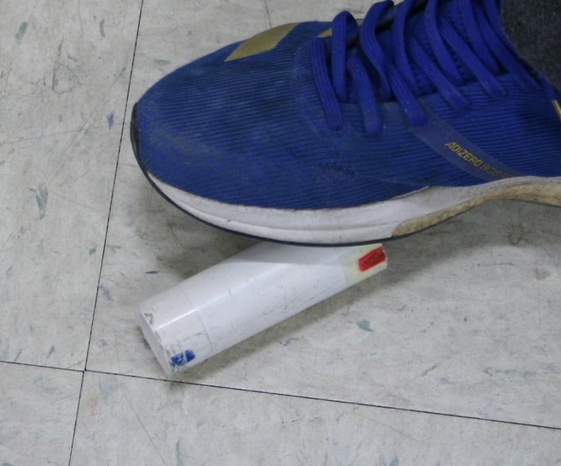 Blue sneaker resting on one end of a white PVC cylinder on a tile floor, demonstrating the foot-squeeze launch technique