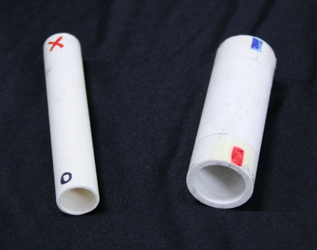 Two white PVC cylinders on dark fabric, each with different colored marks on their ends — left cylinder has a red X on one end and a blue circle on the other, right cylinder has a blue square on one end and a red rectangle on the other