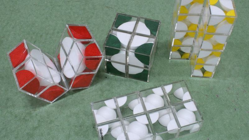 Five transparent folding cube models on green surface with styrofoam sphere sections inside: red-and-white BCC (partially folded), green-and-white FCC (cubic), yellow-and-white NaCl, all-white diamond, and a second white model folded flat