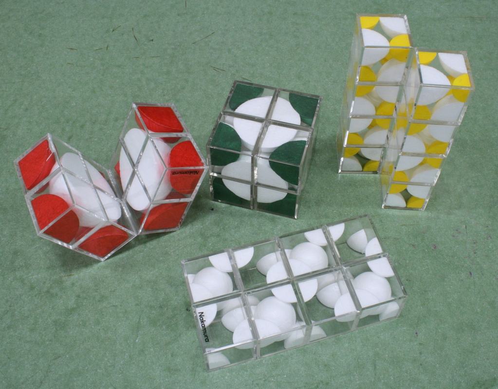 Five transparent folding cube models on green surface with styrofoam sphere sections inside: red-and-white BCC (partially folded), green-and-white FCC (cubic), yellow-and-white NaCl, all-white diamond, and a second white model folded flat