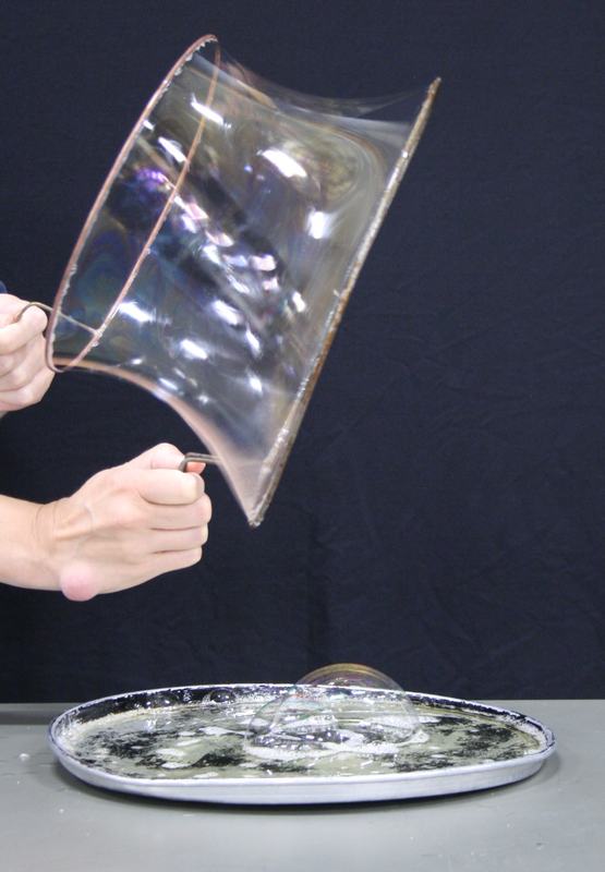 Two hands holding apart two wire rings with a soap film catenoid stretched between them, above a round metal tray of soap solution on a dark background