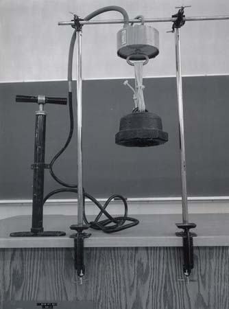 Vacuum cylinder mounted vertically between two support rods with table clamps, hand vacuum pump on left connected by hose, heavy weight hanging from piston