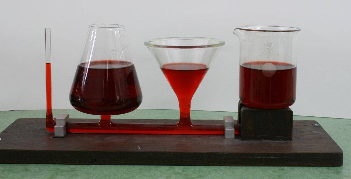Four connected glass vessels—narrow tube, Erlenmeyer flask, funnel, and beaker—mounted on a wooden base and connected by red tubing, all filled with red-colored water to the same height
