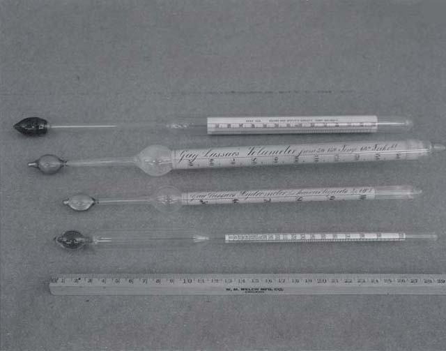 Five glass hydrometers of various sizes laid out on a surface with a ruler for scale