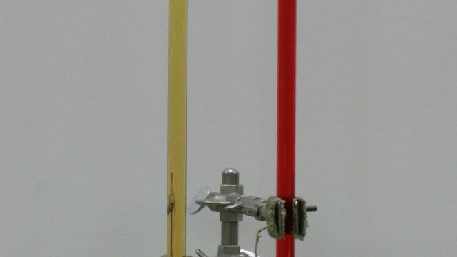U-tube mounted on a ring stand with clamps. Left arm contains yellow oil, right arm contains red-dyed water. The oil column is visibly taller than the water column. The U-bend at bottom is filled with red water.
