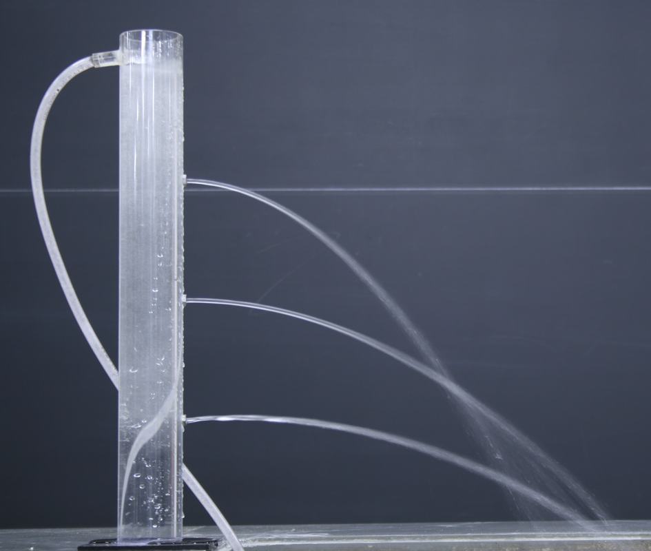 Clear acrylic cylinder filled with water, supply tube entering from the left near the top, three water jets emerging from holes at different heights — lower jets arc farther from the tank