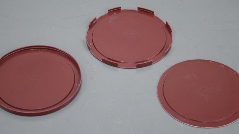 Three dark red plastic coffee can lids on a white surface: left has intact rim, center has partial rim with tab cutouts, right has rim fully removed