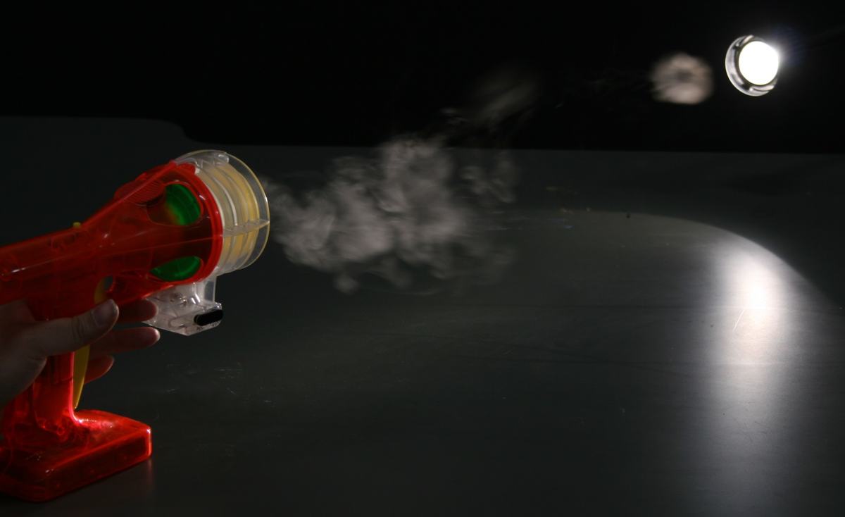Zero Blaster toy firing a smoke ring in a darkened room, illuminated by a spotlight from above