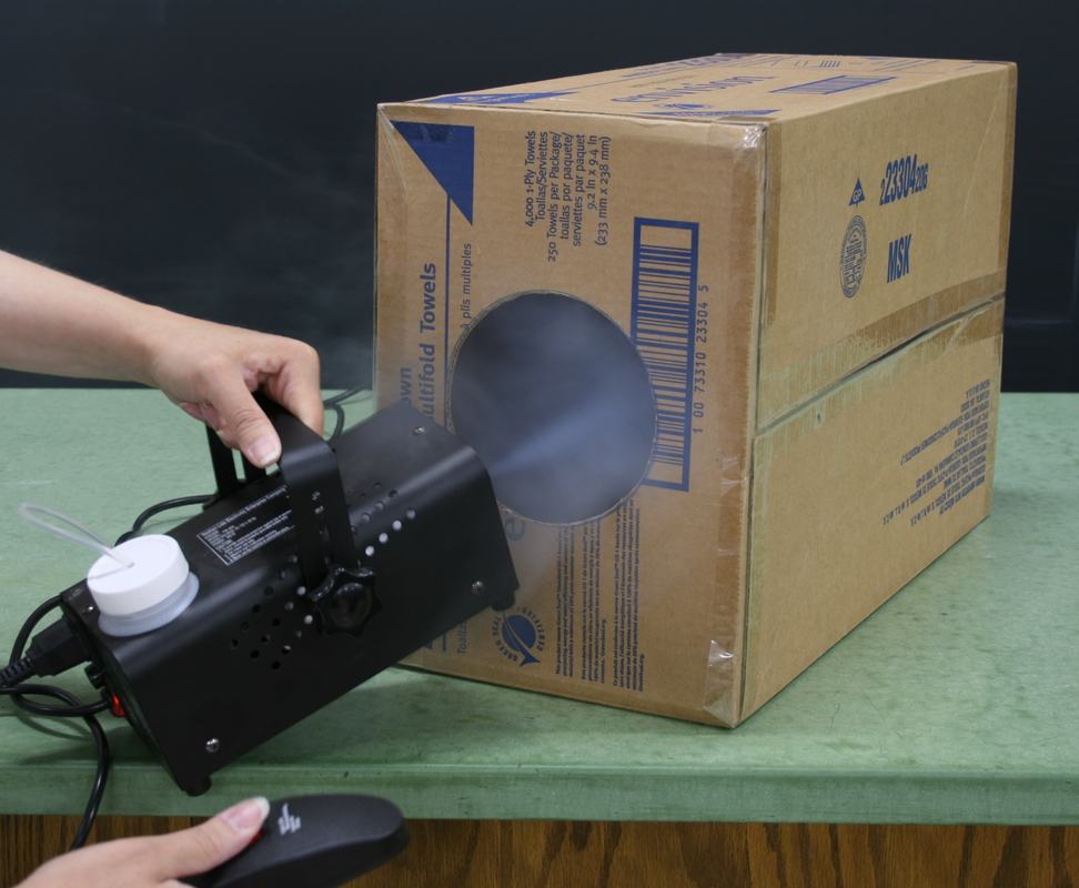 Person directing fog machine output into the circular hole of the cardboard box