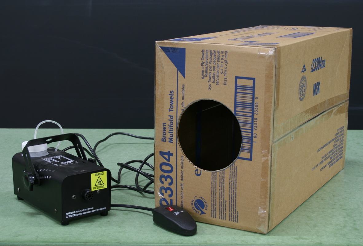 Cardboard box with circular hole cut in one side, fog machine, and wired remote control on a green tabletop