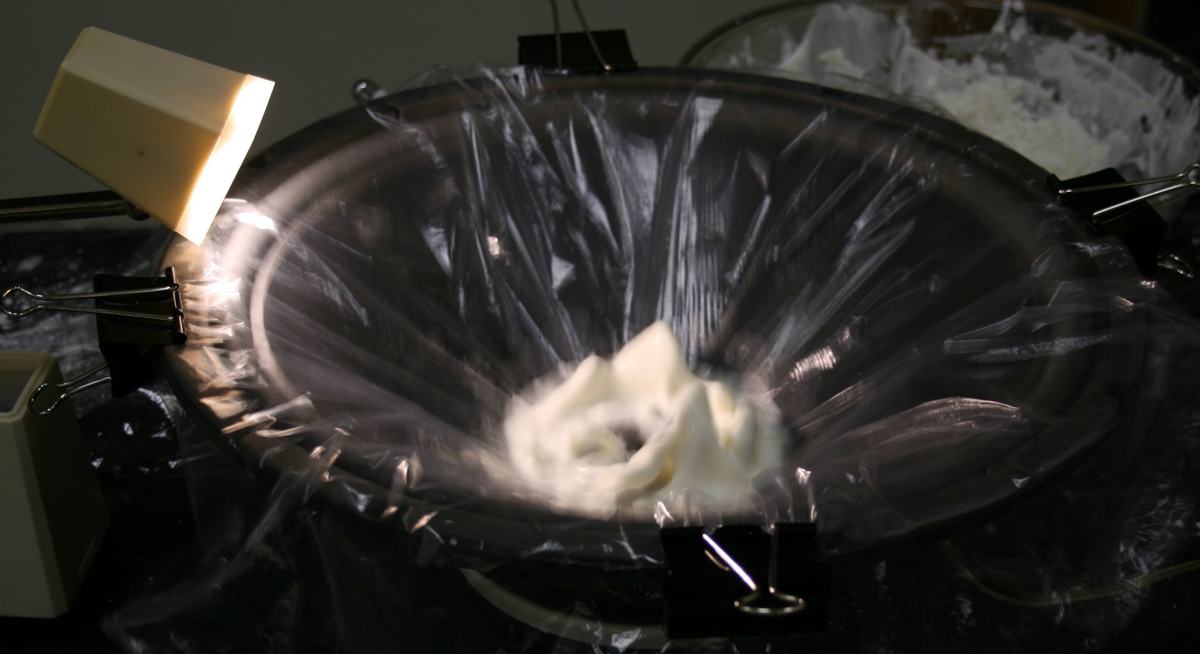 White cornstarch-water mixture forming peaked tentacle structures on a large speaker cone covered with clear plastic wrap, secured with binder clips, illuminated by a small desk lamp