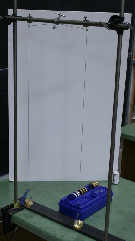 Two pendulums hanging from a horizontal rod between two tall bench clamp stands, with black nylon string looped over ring clamp hooks and brass mass hangers at the bottom; a blue PASCO mass set sits on the bench