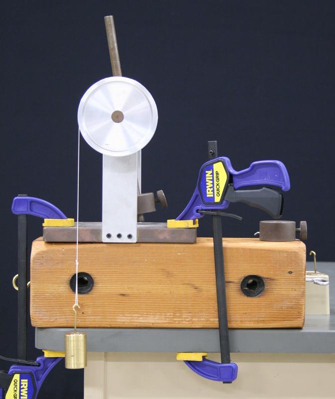 Angular acceleration machine with aluminum disc and metal arm mounted on a wooden block, secured to table with Irwin Quick-Grip clamps; a string from the axle suspends a brass mass