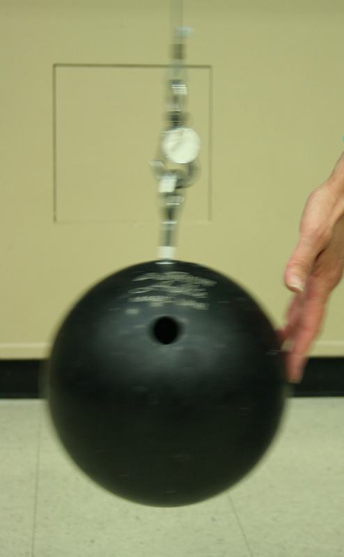 Black bowling ball suspended by cable and hardware, with a hand giving it a gentle push; white floor and yellow wall visible in background