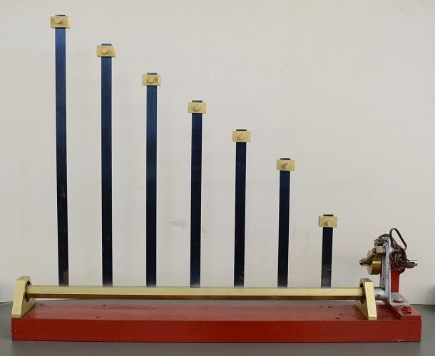 Seven dark blue metal leaf springs of decreasing height with brass block caps, mounted on a brass rail on a red wooden base, with motor and pulley assembly at right end