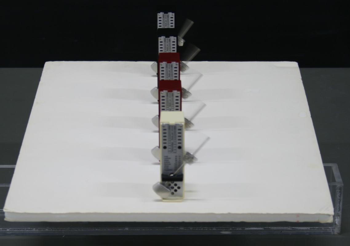 Angled top view of five metronomes running on the foam board, pendulums at different phases