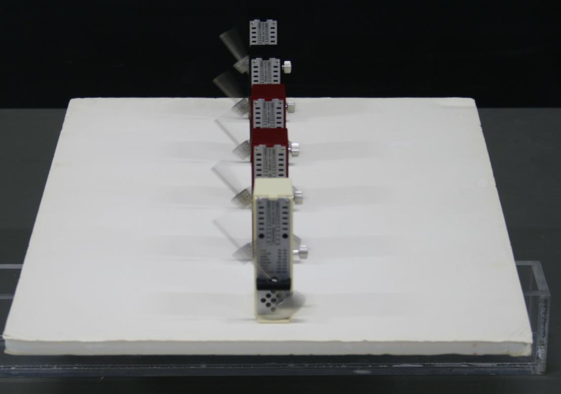 Top view of five metronomes in a line on a white foam board, with pendulums visible swinging perpendicular to the board's rolling direction