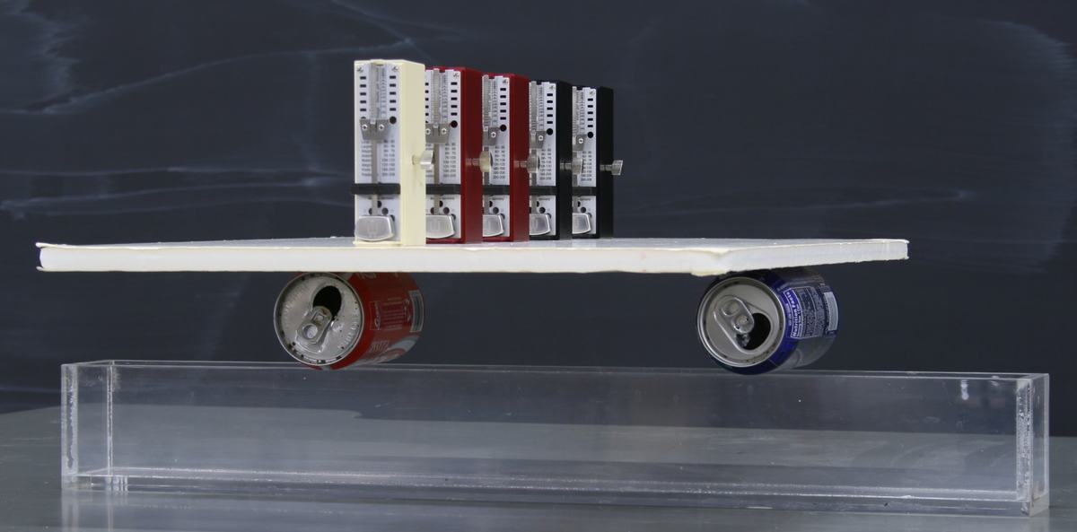 Front view: five metronomes in a row on a white foam board balanced on two soda cans inside a clear plastic tank