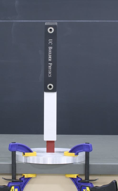 Double pendulum at rest: black upper bar labeled "UC Boulder Physics" with silver lower bar hanging vertically, mounted on a red support column clamped to the table
