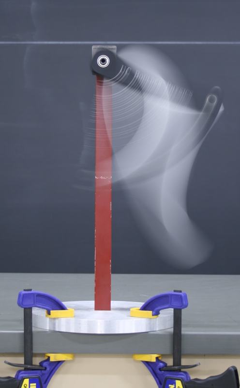 Double pendulum in motion: a red vertical support bar topped by a black pivot bracket, with a blurred silver bar swinging through a wide arc. The base is clamped to the table with blue and yellow clamps.