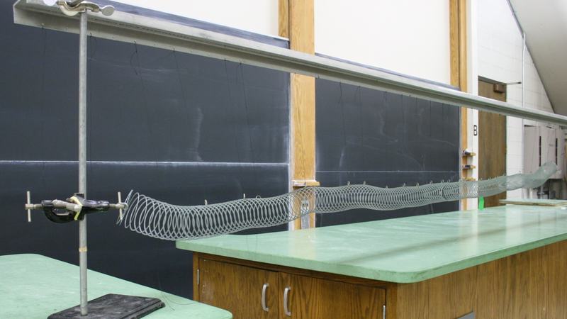 Metal slinky suspended by strings from a horizontal rod between two ring stands on a lecture bench, with one end clamped to the stand
