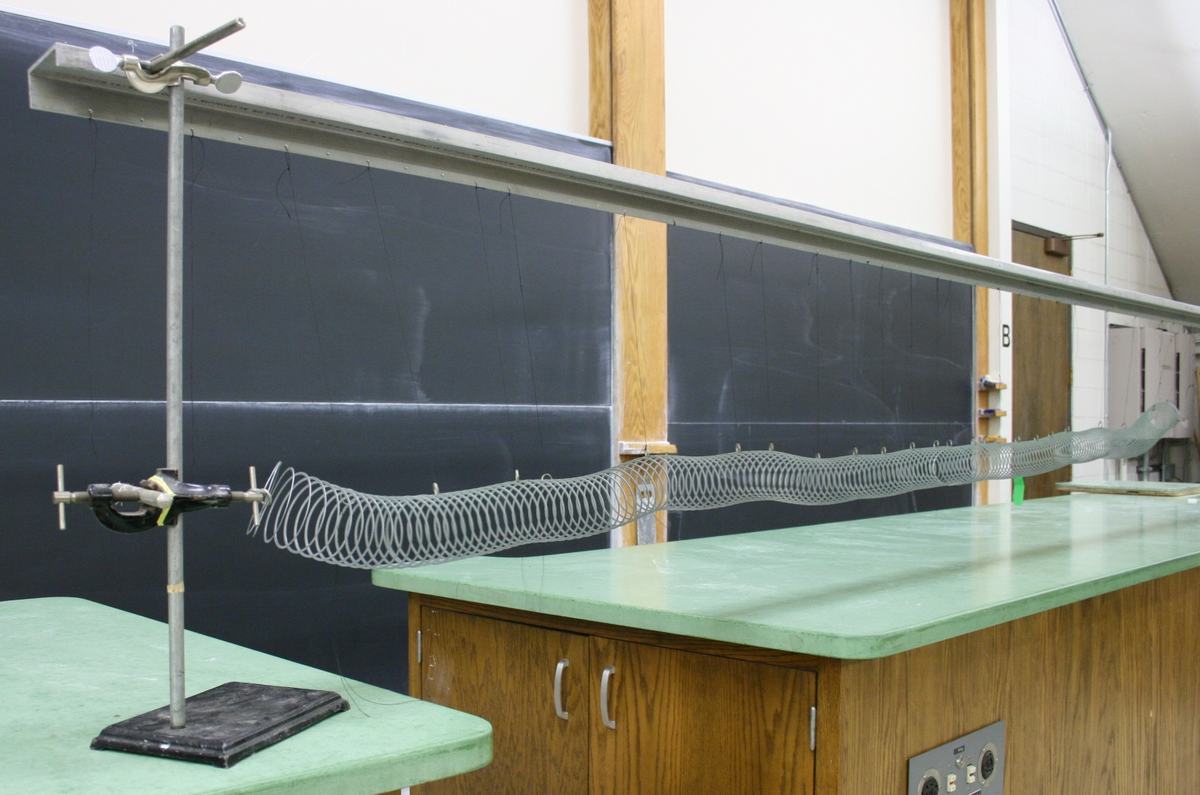 Metal slinky suspended by strings from a horizontal rod between two ring stands on a lecture bench, with one end clamped to the stand