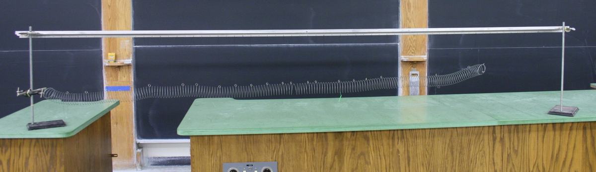 Full view of the suspended slinky spanning the lecture bench between two ring stands, with the slinky hanging from strings attached to a horizontal rod