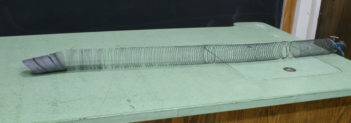 Large metal Slinky stretched along a green lecture bench, clamped at the far end, with compressed coils visible at the near end