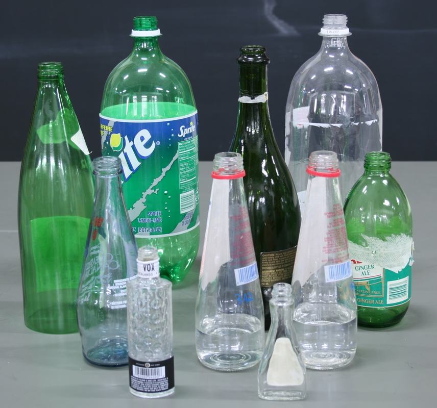 Collection of about ten bottles of various sizes — a large plastic Sprite bottle, green glass bottles, a dark champagne-style bottle, and several clear glass and plastic bottles arranged on a gray surface