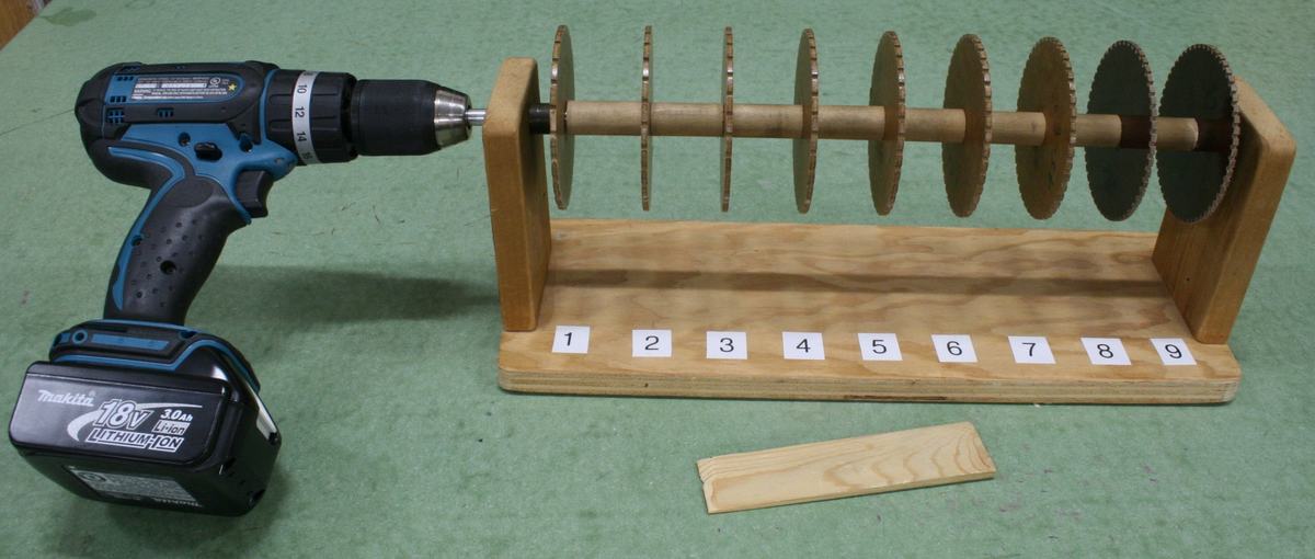 Nine toothed disks mounted on a horizontal shaft in a wooden frame with numbered positions 1-9, a blue cordless drill at the left end, and a wooden shim in front