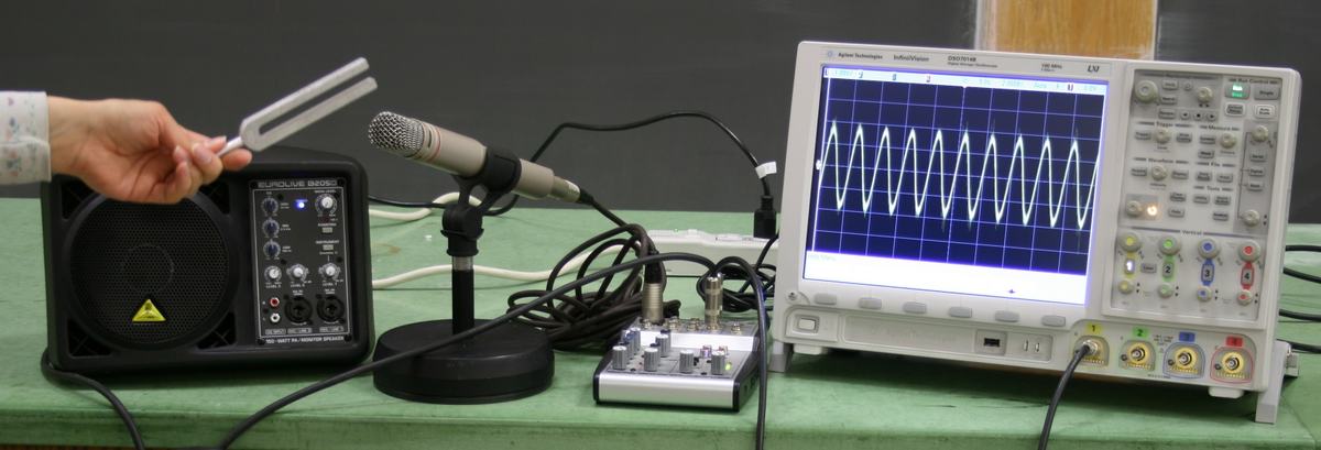 Agilent InfiniiVision oscilloscope displaying a sinusoidal waveform from a tuning fork; AKG microphone on desk stand, Behringer powered monitor speaker, and audio adapter box on table; hand holding tuning fork near microphone