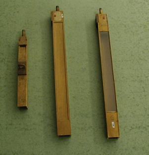 Three wooden organ pipes of increasing length lying on a green surface; the smallest has a removable stop, the middle one has a thin wood membrane panel, and the longest has a glass window