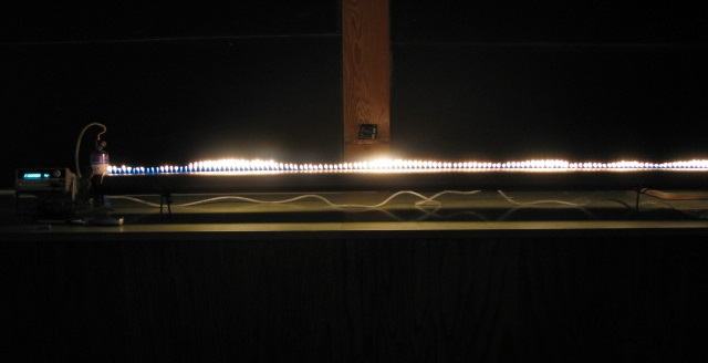 Ruben's tube in a darkened lecture hall showing a row of small flames along the full length of the copper tube, with speaker and signal generator at the left end
